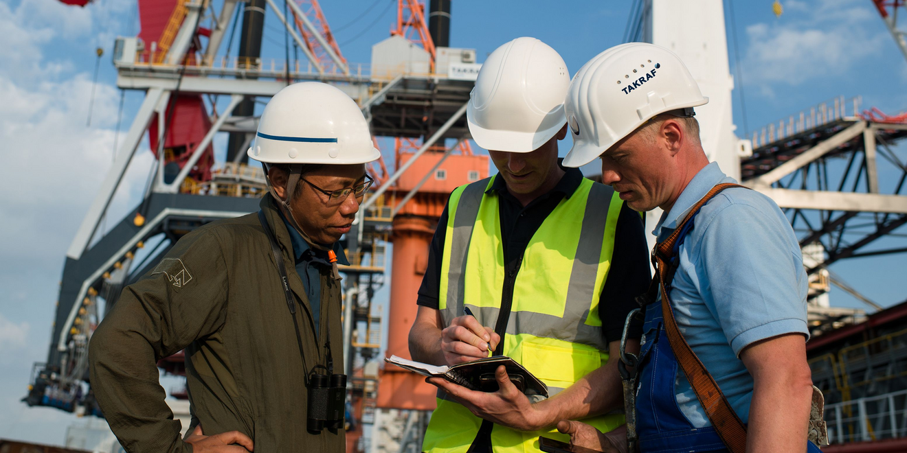Three men wearing white safety helmets and high‑visibility vests stand together at an industrial site, examining plans or equipment as heavy machinery and steel structures loom in the background.