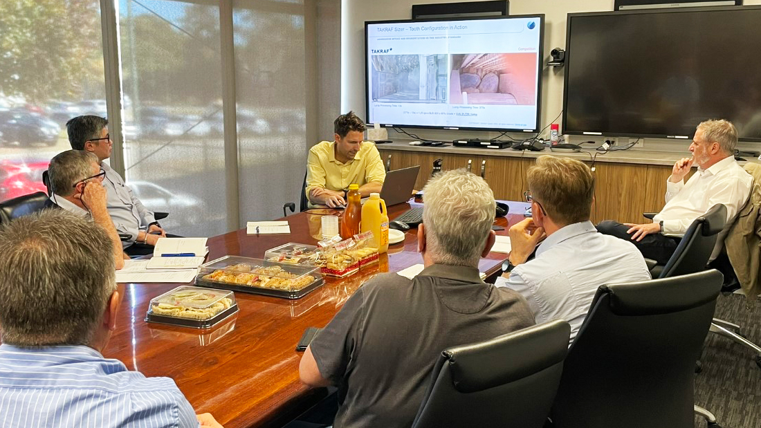 Seven men sitting around a conference table with food in the middle, one man on his laptop showing a presentation